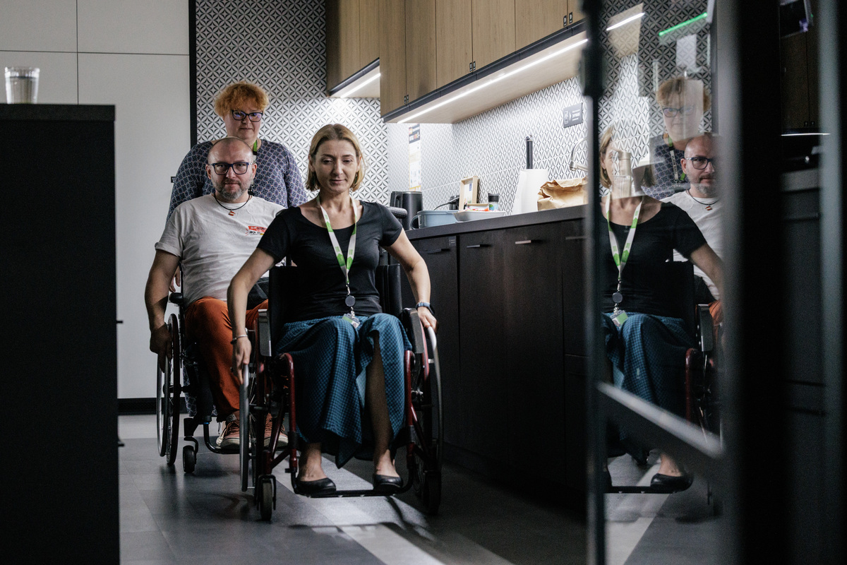 Uczestniczka siedzi na wózku i porusza się po kuchennej części biura, a za nią jedzie druga osoba na wózku. Obok stoi osoba wspierająca ćwiczenie. Zadanie pokazuje codzienne funkcjonowanie i mobilność w przestrzeni pracy.